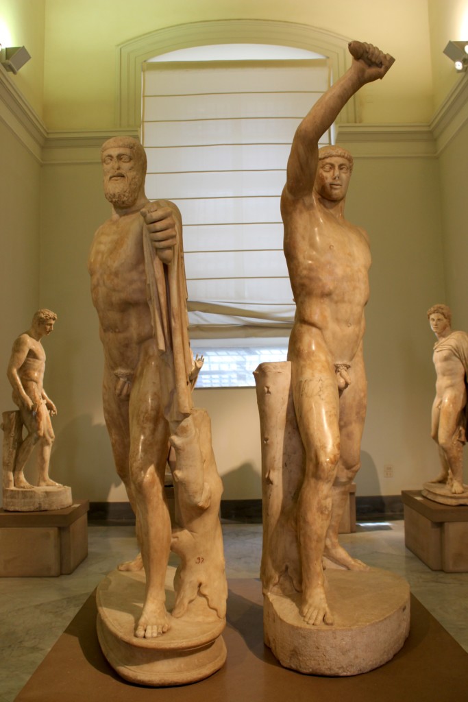 Two ancient marble statues stand upright in a museum, one with a raised arm and the other holding a draped garment. Additional statues are visible in the softly lit room.