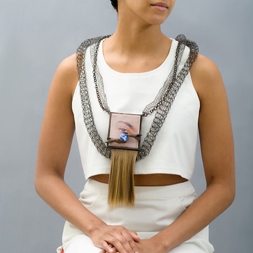 A person wears an intricate necklace featuring a large pendant with an eye image and dangling hair. The necklace is composed of woven chains, set against a plain, light-colored outfit in a neutral setting.