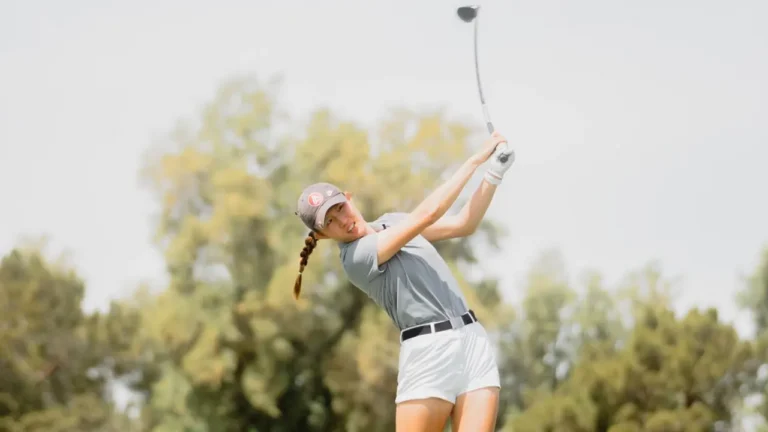 A golfer swings a club on a sunny day, wearing a gray shirt, white shorts, and a cap with a red logo; trees are blurred in the background.