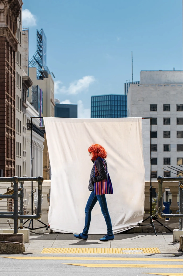 A person with red hair walks past a white backdrop on a city street, surrounded by tall buildings under a clear blue sky.