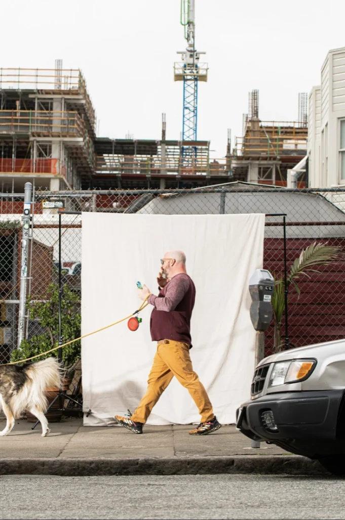 A person in colorful attire walks a dog while using a phone, passing a construction site behind a white backdrop on an urban street. A vehicle is partially visible in the foreground.