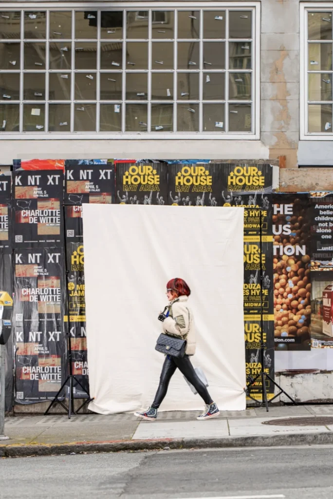 A person walks past a large fabric backdrop set against a wall covered with event posters. Text includes "OUR HOUSE BAY AREA" and "CHARLOTTE DE WITTE." A windowed facade features on top.
