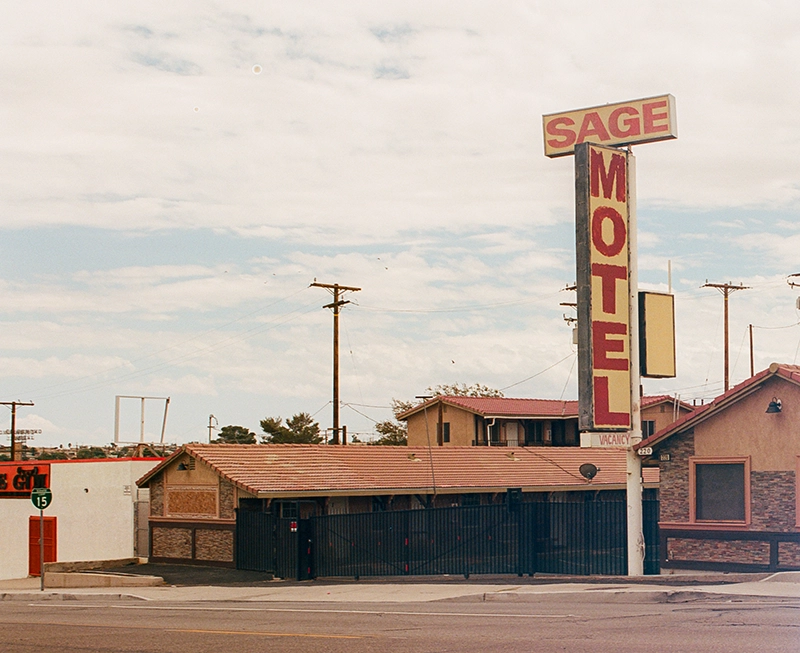 A large sign reading "SAGE MOTEL" stands prominently, indicating vacancy, in front of a modest building with a tiled roof. Utility poles and a clear sky create a roadside setting.