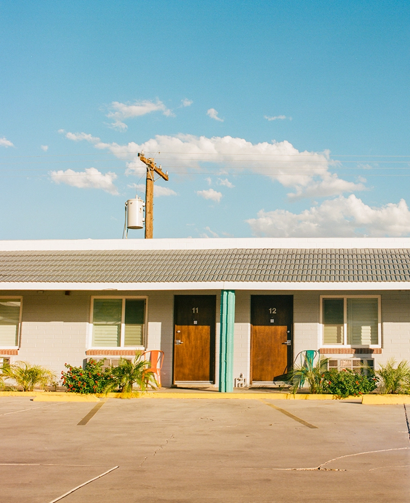 Two wooden doors numbered 11 and 12 sit side by side on a motel building. A clear sky with light clouds and an electrical pole backdrop the scene.