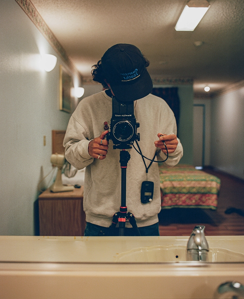 A person photographs themselves in a mirror using a camera on a tripod. They wear a hat and sweatshirt in a room with a bed, lamp, and sink visible.