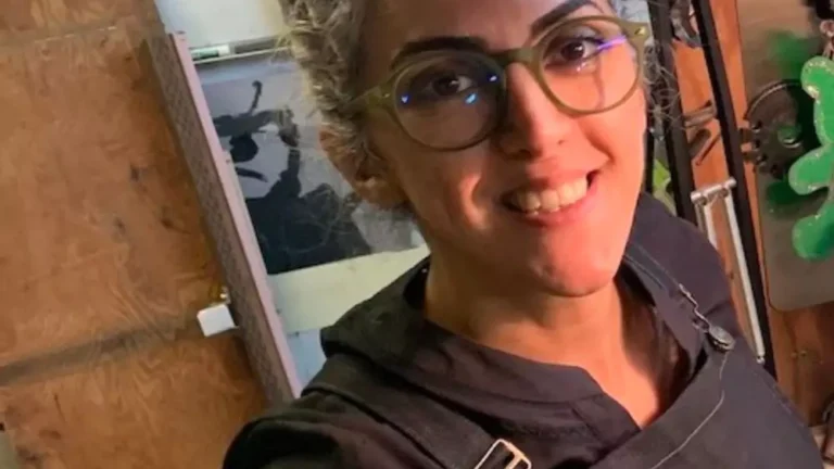 Person smiling, wearing glasses and a dark apron, standing in a workshop. Background includes wooden boards, a hanging tool, and a partially visible whiteboard.