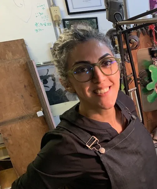 A person with glasses smiles toward the camera, wearing dark clothing and an apron, standing in a cluttered workspace with art supplies and framed art visible in the background.
