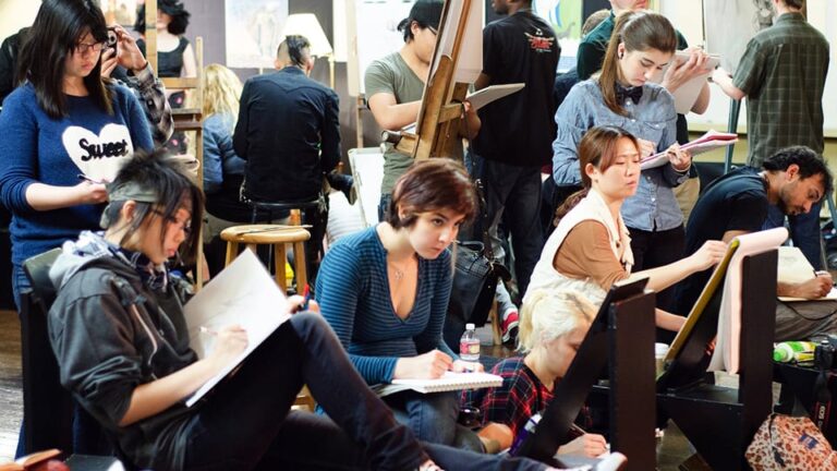 Students draw on sketchpads, concentrating intently in a crowded art studio. An easel and various artworks are visible. One student wears a blue sweater with a white heart and the word 