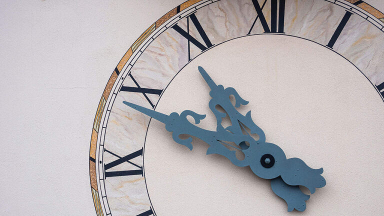 A clock with ornate hands points at Roman numerals on a textured circular face mounted against a plain wall, reflecting a classic architectural design.