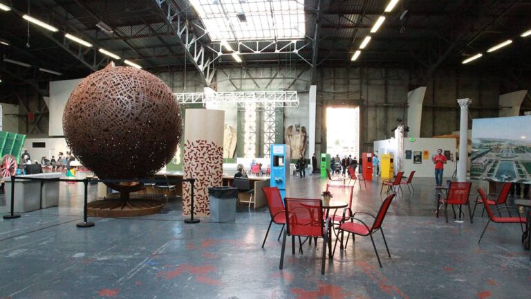 Large sculpted metal sphere displayed indoors; surrounding red chairs and tables on a wide, industrial concrete floor. People and informational panels scattered throughout the spacious, high-ceilinged exhibition hall.