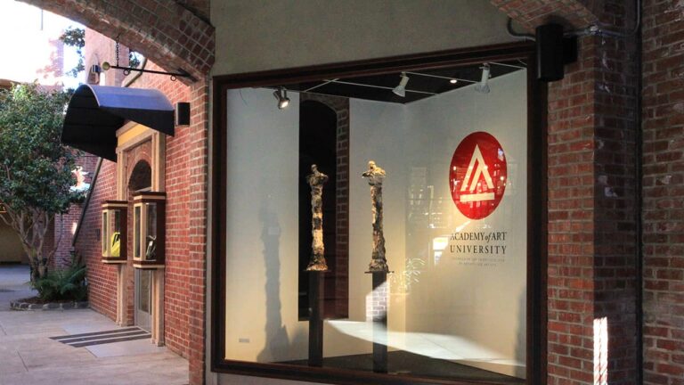 Two tall sculptures stand in a glass display case beside a brick walkway. The window bears the 