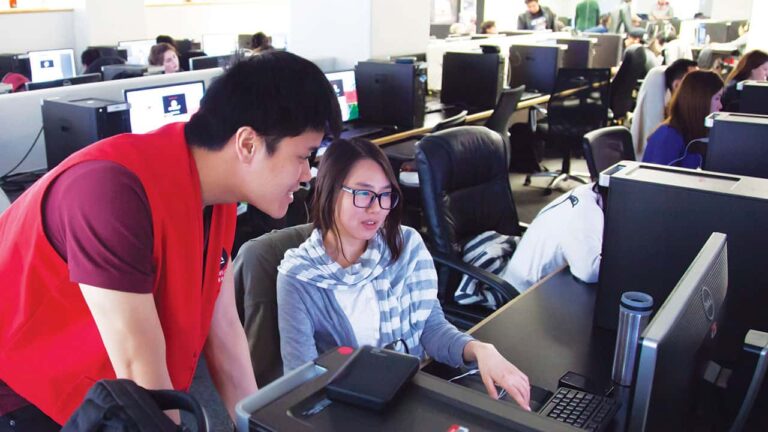 A woman sits at a computer explaining something to a standing man in a red vest in an office environment filled with people working at computers.
