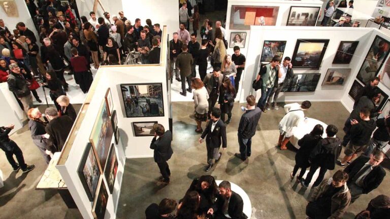 People mingle and observe artwork displayed on walls in a busy art gallery. The space is filled with paintings and visitors, creating a lively, cultural atmosphere.
