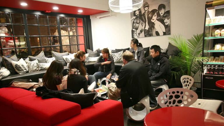 A group of people sit conversing on black and red furniture, surrounded by art, pillows, and plants, in a cozy indoor lounge with a large window and snack shelf.