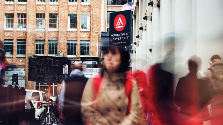 People walk on a busy sidewalk in front of Academy of Art University signage. A brick building and a construction sign with 