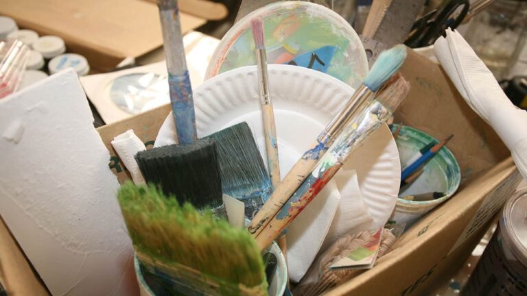 Paintbrushes sit upright in a container, surrounded by paper plates and art supplies within a cluttered studio setting. No text is present.
