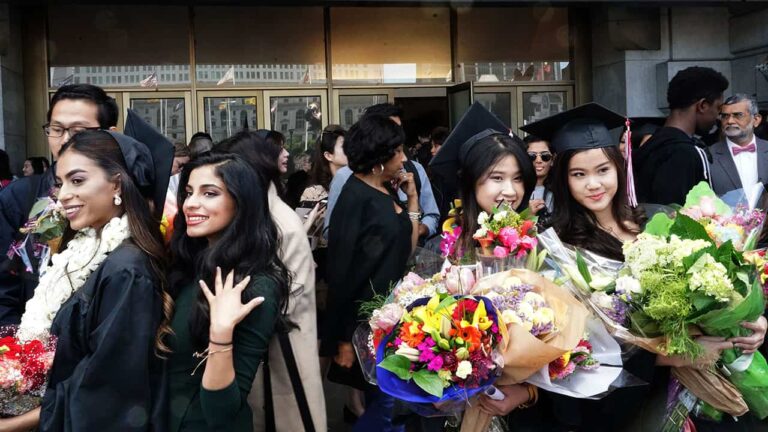 Graduates holding vibrant flower bouquets smile in a crowded outdoor celebration. People, some wearing caps and gowns, mingle near a building entrance.