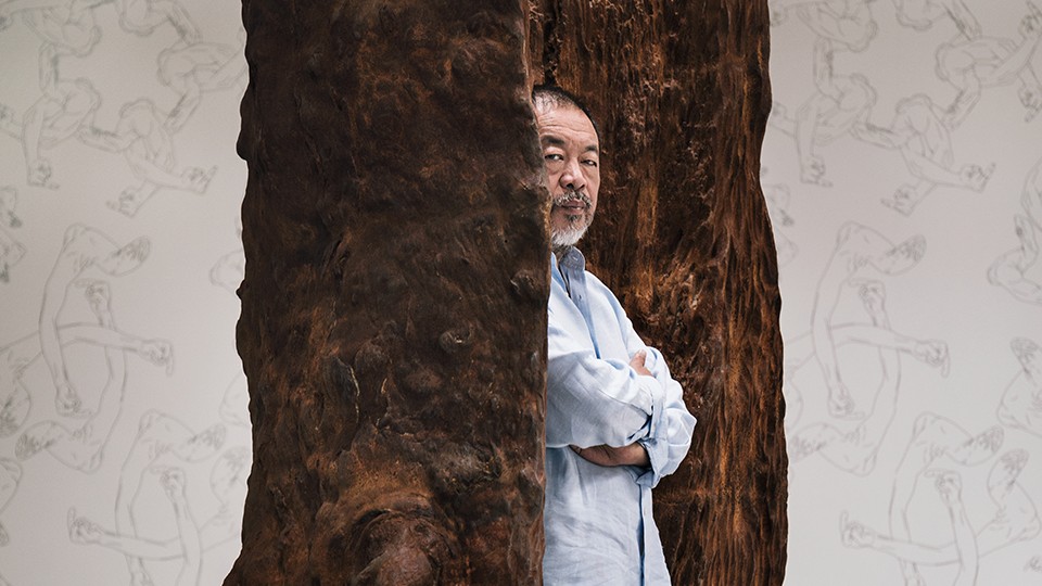 A person stands with folded arms, partially obscured by large, tree-like sculptures, in a room with repeating line-drawings of human figures on the walls.