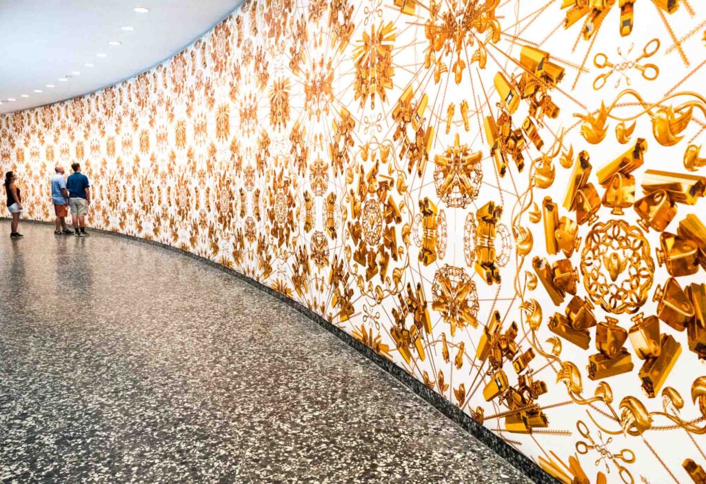 A patterned wall featuring intricate golden designs viewed by three people in a modern, curved gallery with a terrazzo floor.