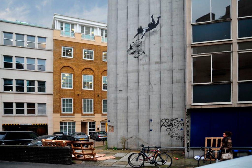 A mural depicts a person falling with a shopping cart on a gray urban building wall. Graffiti text reads, "COPS AGAINST CUTS - The G-20 COPS." Nearby are buildings, cars, and bicycles.