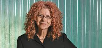 A person with curly hair stands smiling in front of a textured green background, wearing glasses and a black top. No text is visible in the image.