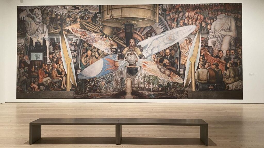 A large mural depicting an industrial scene with a central figure operating machinery, surrounded by crowded figures and intricate details, displayed on a gallery wall above a wooden bench. No text present.