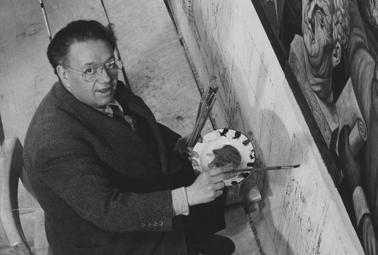 A person holds a paintbrush and palette while painting a mural on a vertical surface, seated on a wooden platform, wearing glasses and a suit jacket. The mural depicts intricate figures.