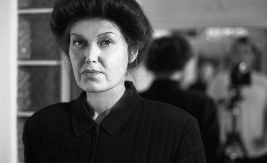 A person stands facing forward in a dimly lit, possibly vintage room with mirrors and a camera in the background, wearing a dark, textured coat.