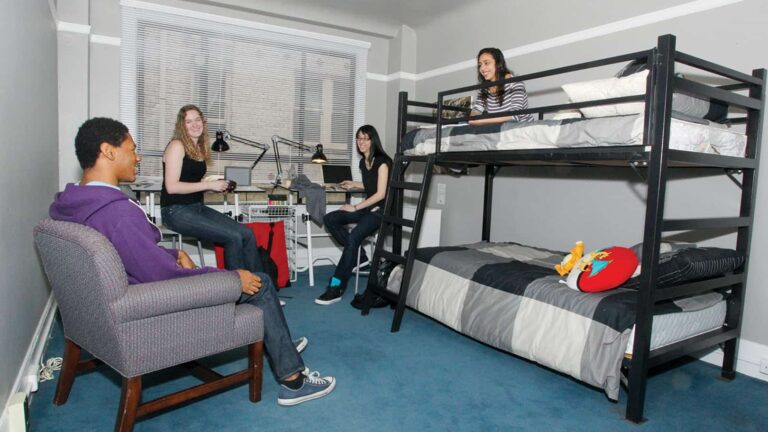 Four people sit and chat in a dorm room with a bunk bed, desk, and chair. Laptops and lamps are on the desk. Room has blue carpet and gray walls.