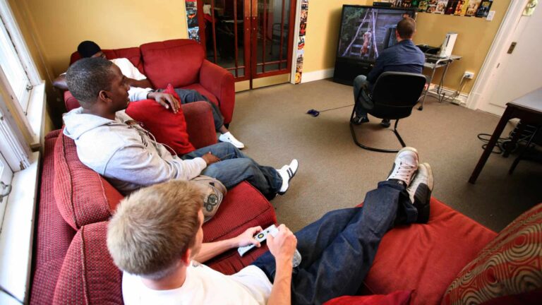 Four people relax in a living room; three sit on red couches, watching the fourth play a video game on a large TV. The room's walls display posters.