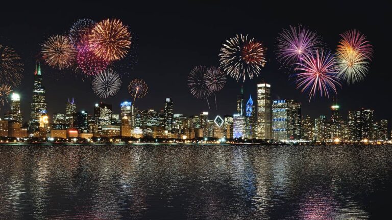 Fireworks burst above a brightly lit city skyline at night, reflecting on a calm body of water, creating a vibrant and festive atmosphere.