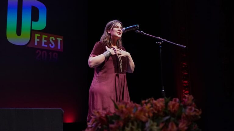 A person stands on stage speaking into a microphone, hands on chest. The backdrop reads 