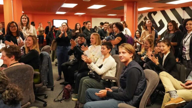 Audience claps enthusiastically in a brightly lit room with orange walls and office chairs, suggesting a celebratory or congratulatory event at an art and design university.
