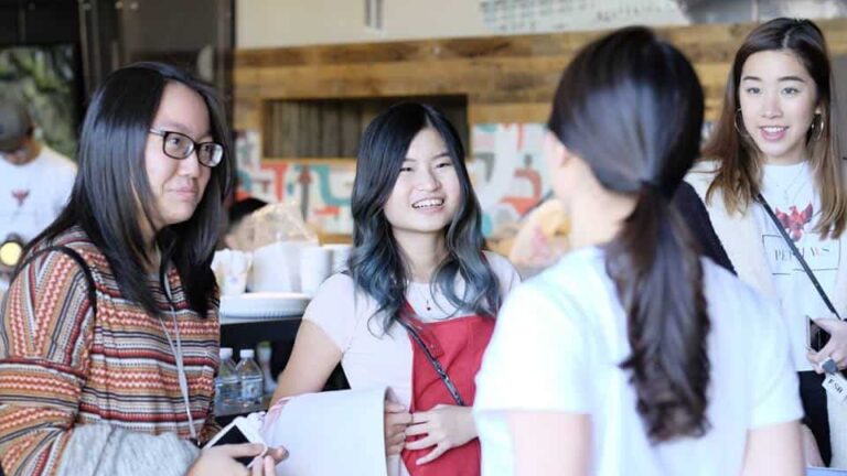 Four individuals stand together, smiling and conversing indoors. Background features abstract art and wooden accents, suggesting a casual, creative environment. One holds papers, another wears a striped sweater.