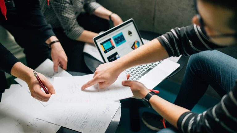 Group of people collaborating, examining papers with handwritten notes and a laptop displaying colorful designs, seated at a table in a modern workspace.