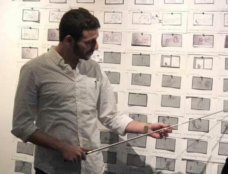 Man holding a pointer gestures towards storyboard pinned on a wall. He is wearing a patterned shirt, focused on explaining visual sketches and illustrations in an educational setting.